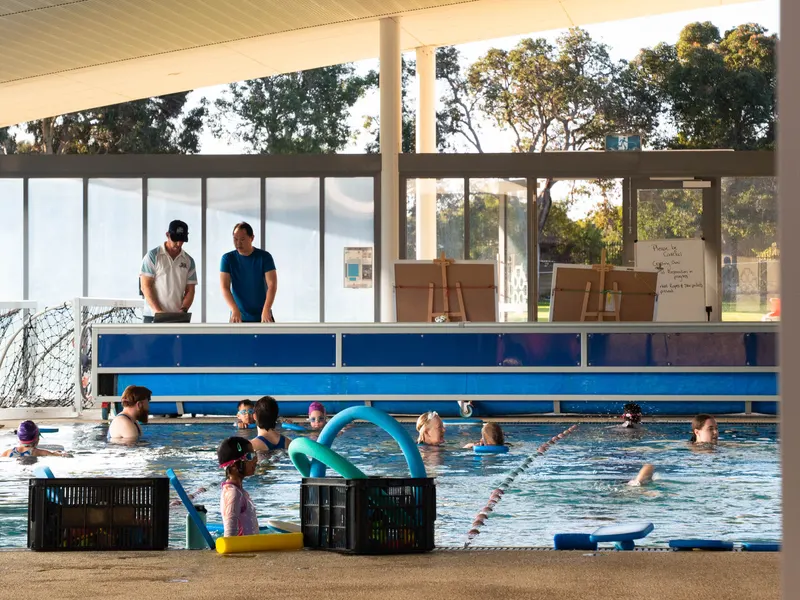 The outdoor Learn to Swim pool during a lesson, with children in the shallow water using kick-boards and pool noodles while two instructors watch from the deck