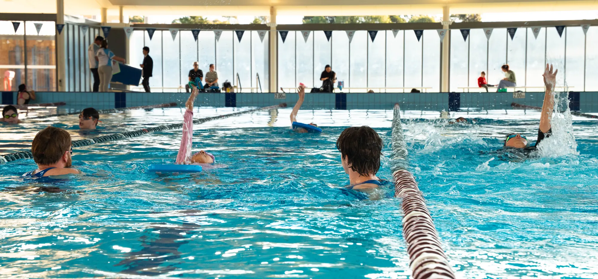 A backstroke class mid-lesson in the heated pool at Corpus Christi College Aquatic Centre, with an instructor in the water alongside the swimmers