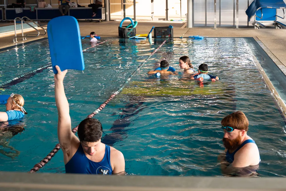Instructors in the shallow heated pool running a kickboard drill with young children
